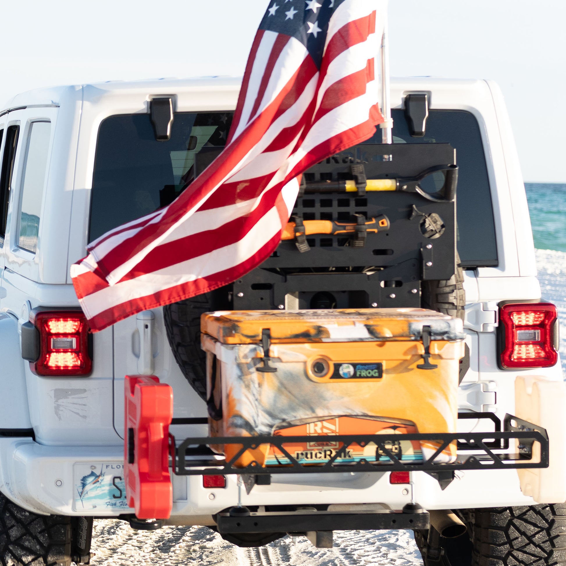 Back of a white SUV with a cooler and American flag, parked outdoors.
